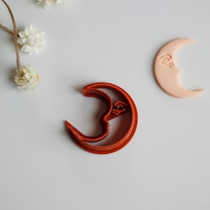 May include: Two crescent moon cookie cutters, one red and one light pink. The red cookie cutter is a double-sided design with a raised detail on the inside of the crescent.