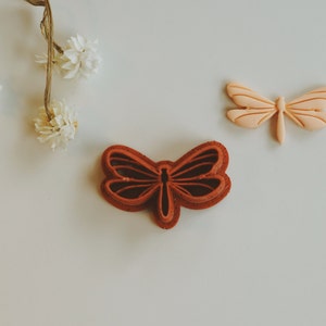May include: Two dragonfly cookie cutters, one in a light peach color and one in a dark red color. The cutters are on a white surface.
