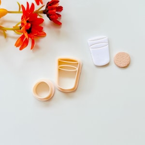 May include: A collection of polymer clay cutters in the shape of a glass, a coaster, and a cup. The glass cutter is light peach, and the coaster is a light tan color. A white cup-shaped cutter is also present. A sprig of orange flowers is in the upper left corner.