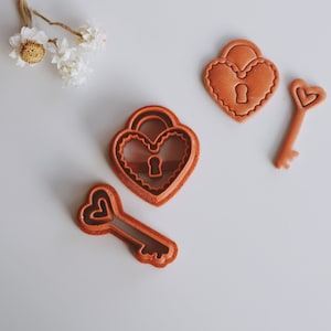 Op de afbeelding: Vier oranje hartvormige koekjessnijders worden getoond op een witte ondergrond. De set bevat twee sloten en twee sleutels, elk met ingewikkelde details. De sloten hebben sleutelgaten en de sleutels hebben hartvormige uitsparingen. Gedroogde bloemen zijn op de achtergrond.
