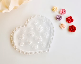 May include: A clear, heart-shaped mould featuring rose designs, with a scalloped edge. Several small, colourful rose-shaped embellishments in red, pink, purple, and yellow are scattered nearby. Suitable for crafting and DIY projects.