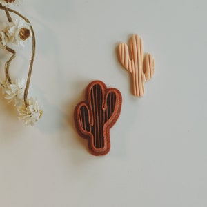 May include: Two cactus-shaped cookie cutters, one brown and one light peach, on a white surface.