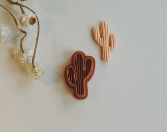 Peut inclure: Deux emportes-pièces en forme de cactus, l'un brun et l'autre pêche clair, sur une surface blanche.
