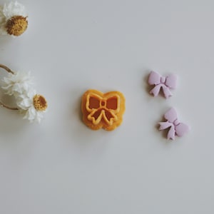 Op de afbeelding: Drie kleine decoraties in de vorm van een strik. Eén is een lichtoranje strik met een uitgesneden ontwerp. De andere twee zijn lichtpaars.