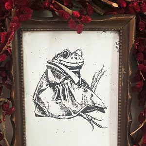 Puede incluir: Una ilustración en blanco y negro enmarcada de una rana con una chaqueta. El marco es de madera marrón con un borde decorativo. El fondo es una corona de bayas rojas.