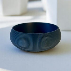 May include: A small, round, dark blue bowl with vertical ribbed texture on the exterior. The interior is a darker shade of blue. The bowl sits on a white surface, with white geometric planters and succulents in the background.