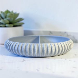 May include: A low-profile, circular light grey tray with a textured, ribbed exterior and a single interior divider that splits the basin into two equal sections. The surface of the tray has a speckled, matte finish throughout its entire structure.