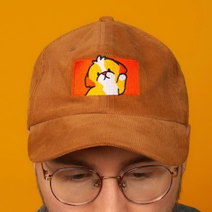 May include: A brown corduroy baseball cap with an embroidered patch of a cartoon dog with a yellow body, white face, and orange ears. The dog is wearing a red collar and has a white paw raised in the air.