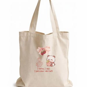 May include: A cream-colored canvas tote bag featuring a graphic print of a pink ribbon with two strawberries, a small panda holding a strawberry, and two strawberry outlines. The text reads: I came, I saw. I panicked and left.