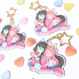 Peut inclure: Deux personnages de dessin animé, une princesse aux cheveux roses et une fille aux cheveux noirs, se tiennent la main et sourient. Elles portent toutes les deux des tenues avec des cœurs et des étoiles. Les personnages sont sur un porte-clés en acrylique transparent avec un charme d'étoile dorée.