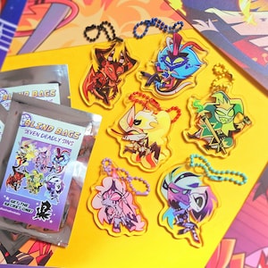 Puede incluir: Una colección de llaveros acrílicos coloridos con personajes de dibujos animados y varias "Blind Bags" selladas con la inscripción "Seven Deadly Sins". Los llaveros tienen cadenas de cuentas. El fondo es amarillo y morado.