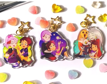 May include: Three colorful acrylic keychains featuring cartoon characters from the animated series The Owl House. Each keychain features a different couple from the show: Luz and Amity, Hunter and Willow, and Eda and King. The keychains are attached to gold-colored star-shaped charms.