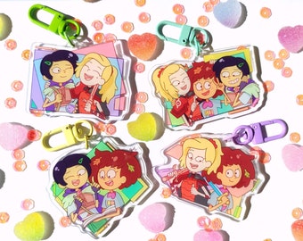 Könnte beinhalten: Vier bunte Schlüsselanhänger mit Cartoon-Charakteren, die drei Mädchen mit braunen Haaren und ein Mädchen mit blonden Haaren zeigen. Die Figuren tragen verschiedene Outfits und halten Bücher. Die Schlüsselanhänger sind aus klarem Acryl gefertigt und haben einen Metallring zum Anbringen an Schlüsseln.