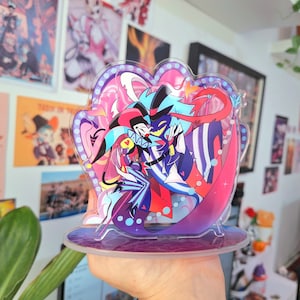 Puede incluir: Un soporte acrílico colorido con dos personajes de dibujos animados, uno con un traje rojo y negro y el otro con un traje azul y blanco, sobre un fondo rosa y azul.