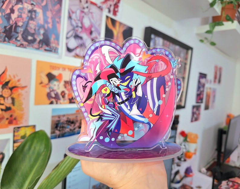 May include: A colorful acrylic stand featuring two cartoon characters, one in a red and black suit and the other in a blue and white suit, with a pink and blue background.
