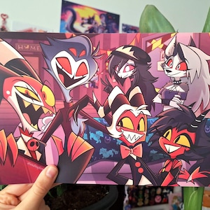 May include: A print featuring six cartoon characters with red, black, and white color schemes. The characters have exaggerated features and are set against a purple and pink background. The print is held by a hand.