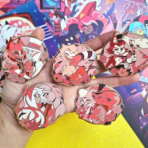 May include: Six round enamel pins featuring cartoon characters from the animated series "Helluva Boss". The pins are gold-colored with red, white, and black accents. The characters are depicted in various poses and expressions.