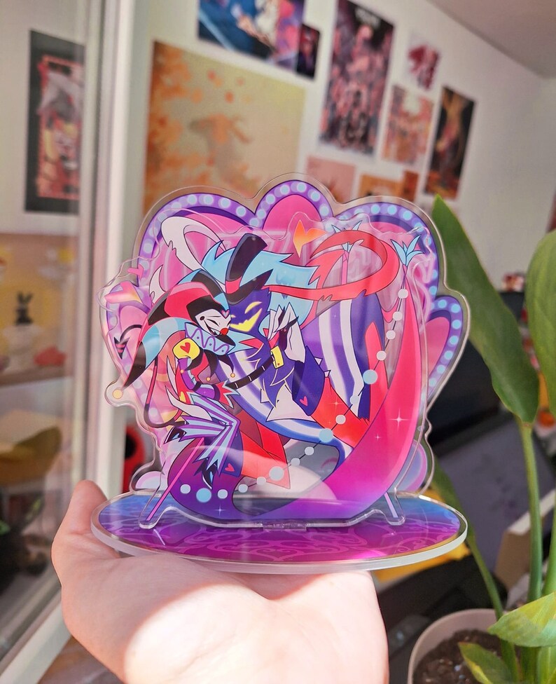 May include: A clear acrylic stand with a colorful illustration of two characters. The characters are a jester with a red and black costume and a blue and purple character with a red bow tie. The stand has a pink and purple base with a white border.
