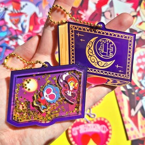 May include: Two purple and gold keychains with a glittery, star-shaped design. One keychain features a crescent moon and a symbol. The other keychain features a cartoon character with a red and blue color scheme.