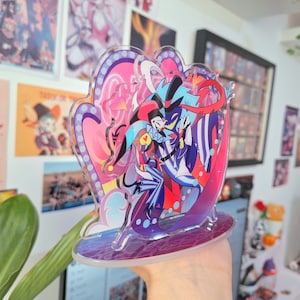 May include: A clear acrylic stand with a colorful illustration of two cartoon characters. The characters are a blue and black jester with a red and white hat and a blue and purple demon with a red horn. The stand has a purple base and a pink and white background.