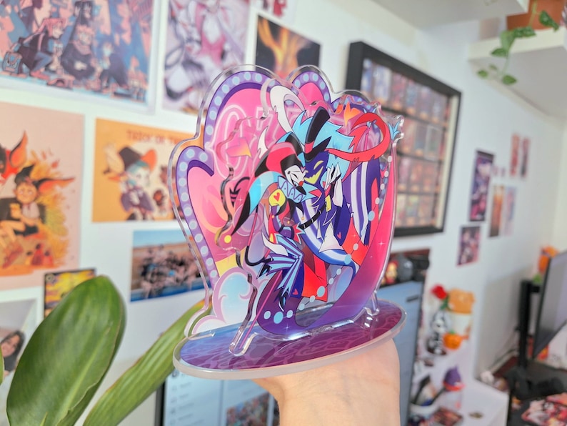 May include: A clear acrylic stand with a colorful illustration of two cartoon characters. The characters are a blue and black jester with a red and white hat and a blue and purple demon with a red horn. The stand has a purple base and a pink and white background.