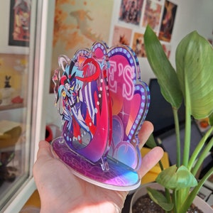 May include: A colorful acrylic stand featuring a character with a jester hat and a red and blue costume. The stand has a pink and purple background with the word "Jester's" in neon pink letters.