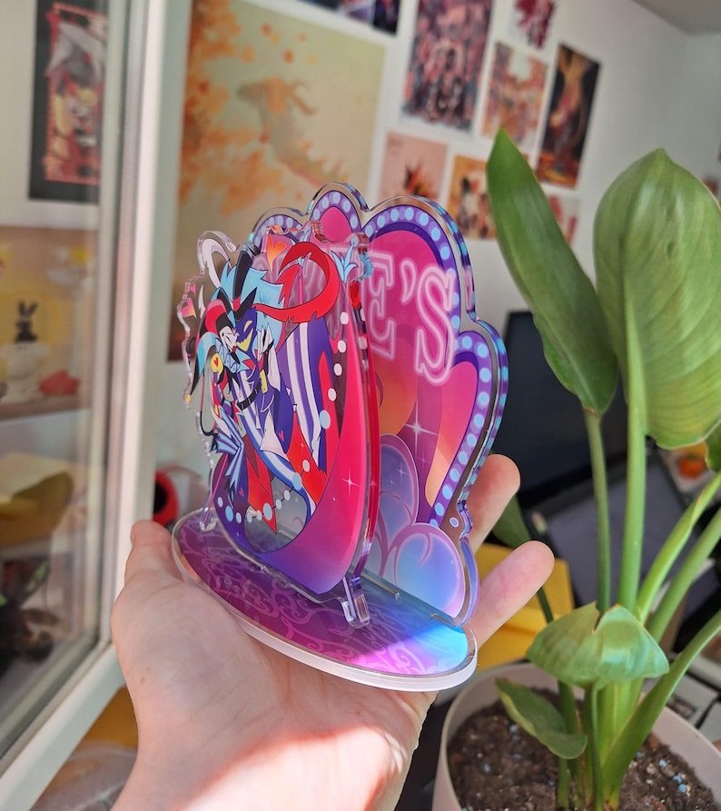 May include: A colorful acrylic stand featuring a character with a jester hat and a red and blue costume. The stand has a pink and purple background with the word "Jester's" in neon pink letters.