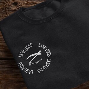 Peut inclure: Sweat-shirt noir avec un motif brodé blanc représentant un œil avec des cils et un outil pour les cils. Le motif est entouré du texte "Lash Boss" répété quatre fois.