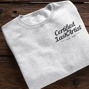 May include: A gray sweatshirt with the text "Certified Lash Artist" in black lettering and a stylized eyelash design.