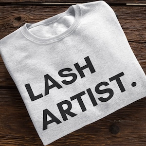 Peut inclure: Sweat-shirt gris avec le texte "LASH ARTIST." imprimé en noir.