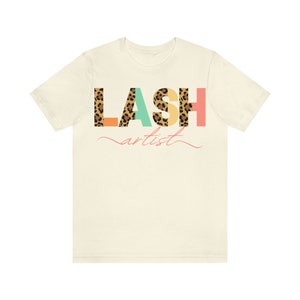 May include: A beige t-shirt with the word "LASH" printed in colorful letters with a leopard print pattern. The word "artist" is written in cursive below the word "LASH".