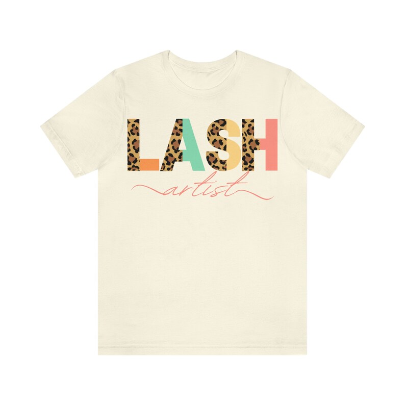 May include: A beige t-shirt with the word "LASH" printed in colorful letters with a leopard print pattern. The word "artist" is written in cursive below the word "LASH".
