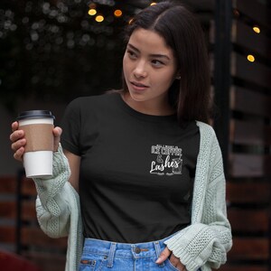 May include: A black t-shirt with white text that reads "All I want is ice coffee & lashes". The text is styled with a coffee cup graphic and eyelashes. A person is wearing the shirt and holding a cup of coffee.