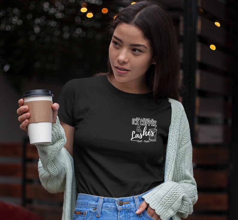 May include: A black t-shirt with white text that reads "All I want is ice coffee & lashes". The text is styled with a coffee cup graphic and eyelashes. A person is wearing the shirt and holding a cup of coffee.