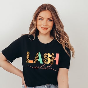 May include: A woman wearing a black t-shirt with the text "LASH artist" in colorful letters with a leopard print design.
