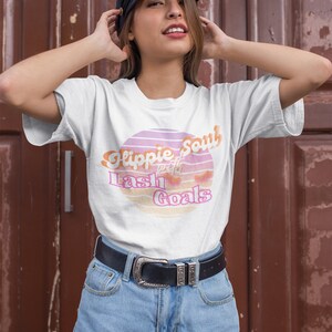 Puede incluir: Una camiseta blanca con un gr&aacute;fico de puesta de sol rosa y naranja que dice "Hippie Soul with Lash Goals". La modelo lleva la camiseta con pantalones cortos de mezclilla y una gorra de golf negra.