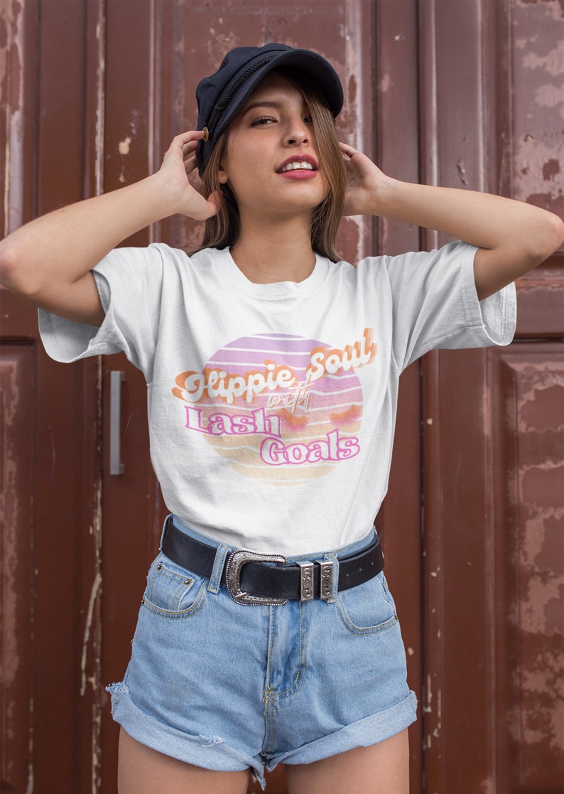 Puede incluir: Una camiseta blanca con un gr&aacute;fico de puesta de sol rosa y naranja que dice "Hippie Soul with Lash Goals". La modelo lleva la camiseta con pantalones cortos de mezclilla y una gorra de golf negra.