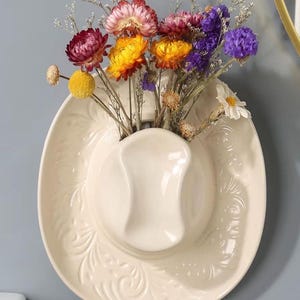 May include: A cream-colored ceramic wall vase shaped like a cowboy hat, filled with a colorful arrangement of dried flowers. The flowers include shades of red, yellow, purple, and white. The hat has embossed floral detailing.