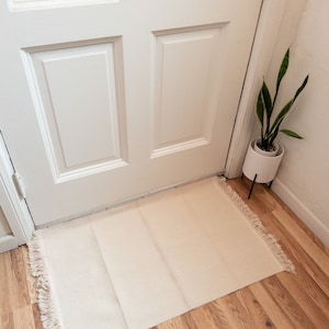 May include: A white doormat with fringe edges sits in front of a white door with a white door knob. The doormat is on a light brown wood floor.