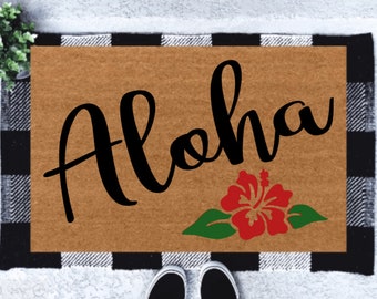 Op de afbeelding: Een bruine deurmat met een zwart-wit geblokte rand. De deurmat heeft het woord "Aloha" in zwarte cursieve letters en een rode hibiscusbloem met groene bladeren.