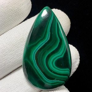 Peut inclure: Une pierre de malachite polie en forme de goutte d'eau. La pierre présente une couleur vert foncé avec des motifs tourbillonnants de teintes vert clair et bleu-vert. Le bijou est tenu par une main gantée sur fond noir.