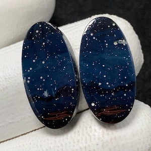 Puede incluir: Dos cabujones ovalados con un diseño de noche estrellada azul oscuro. Puntos blancos representan estrellas contra el fondo azul oscuro. La parte inferior de cada cabujón tiene un color rojo oscuro y marrón, que se asemeja a un horizonte.