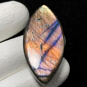 Puede incluir: Una piedra preciosa de labradorita pulida, en forma de marquesa. La piedra muestra una mezcla de colores, incluyendo púrpura, naranja y azul, con un efecto brillante. La gema es sostenida por una mano enguantada sobre un fondo oscuro.