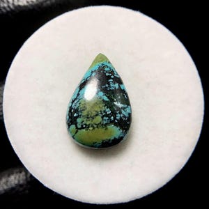 May include: A teardrop-shaped turquoise gemstone with a polished surface. The stone features a mix of blue, green, and black colors, with a speckled pattern. The gemstone is set against a white background.
