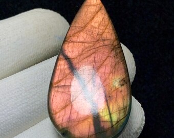 May include: A teardrop-shaped gemstone with a polished surface. The stone displays a blend of pink, purple, and gold hues, with visible internal veining. The gemstone is held against a white fabric background.