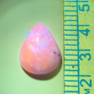 May include: A teardrop-shaped, polished gemstone in shades of pink and orange, with subtle blue and purple inclusions. A yellow and green ruler is positioned next to the gemstone, providing a size reference. The gemstone appears to be approximately 1.5 inches in length.