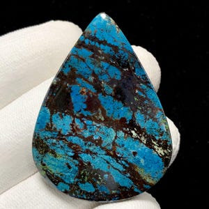 Könnte beinhalten: Ein tropfenförmiger Edelstein mit leuchtend blauer Farbe, durchzogen von dunkelbraunen und schwarzen Mustern. Die polierte Oberfläche reflektiert das Licht und zeigt die natürlichen Farb- und Texturvariationen. Der Stein wird vor weißem Stoff gehalten.