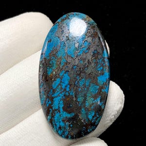 Op de afbeelding: Een ovaalvormige edelsteen met een opvallend patroon van turkooisblauw, donkergrijs en bruin. Het gepolijste oppervlak reflecteert licht, waardoor de ingewikkelde adering en kleurvariaties worden benadrukt. De edelsteen wordt vastgehouden door een gehandschoende hand.