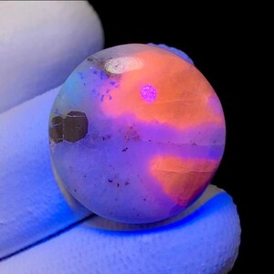 May include: A round, polished gemstone with a vibrant color palette. The stone features a base of pale blue and white, overlaid with patches of orange and purple. A small, bright pink spot is visible on the surface. The gemstone is held by a gloved hand.