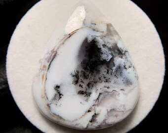 May include: A teardrop-shaped gemstone with a white base, featuring black and gray dendritic inclusions. The stone is set on a white circular surface, with a black background. The gemstone has a polished, translucent appearance.